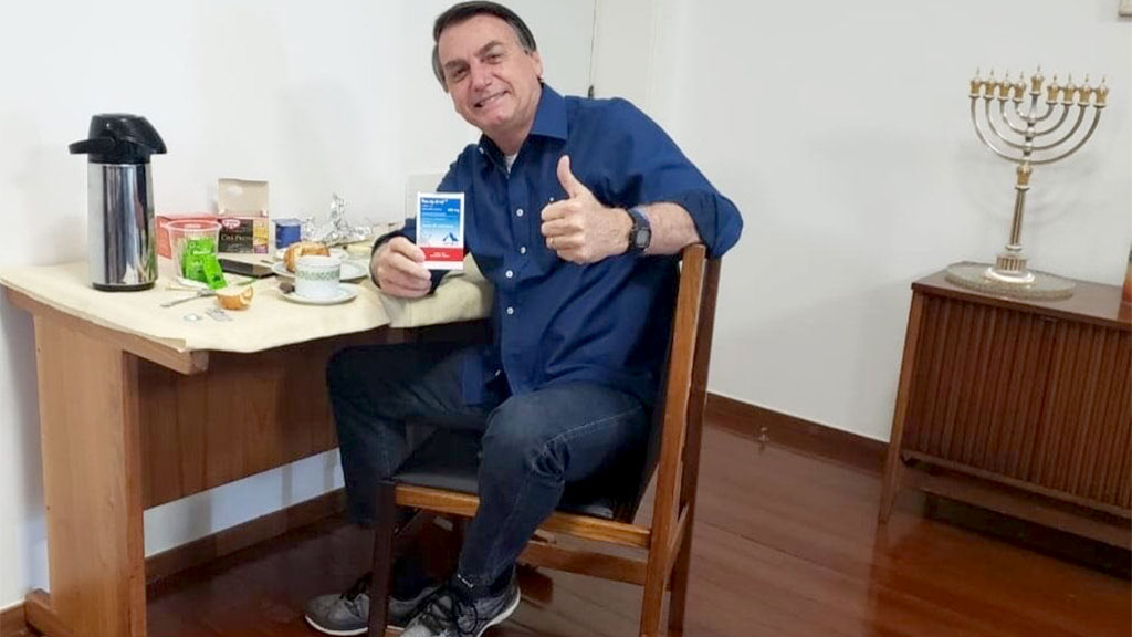 Ao anunciar que estava livre da covid-19, Bolsonaro aparece em um cenário montado com uma caixa de cloroquina na mão e um sinal de positivo